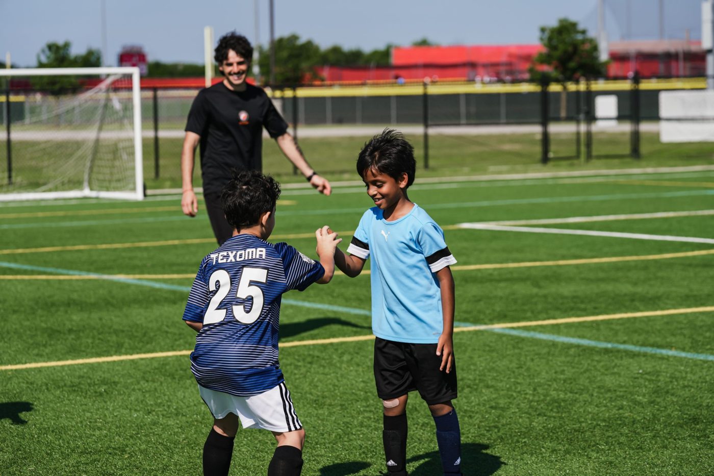 A Foro coach smiles as two young players share a high five on the grass field at The Highlands School